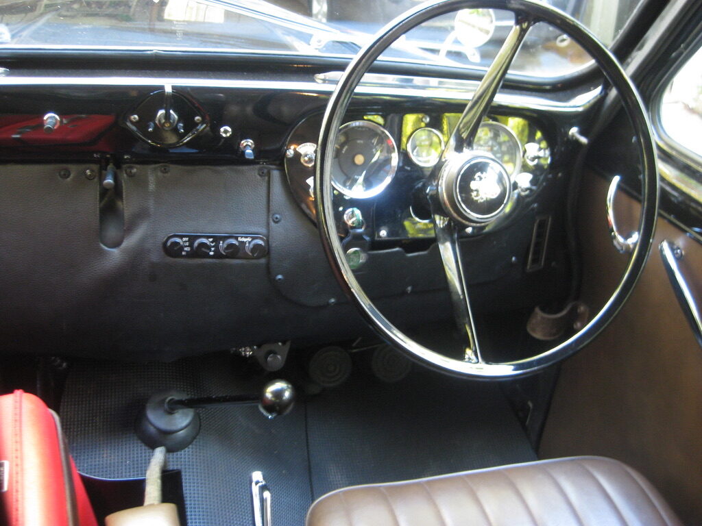 The driver's compartment from the passenger side, right hand drive, large thin rimmed steering wheel and a black painted dash.