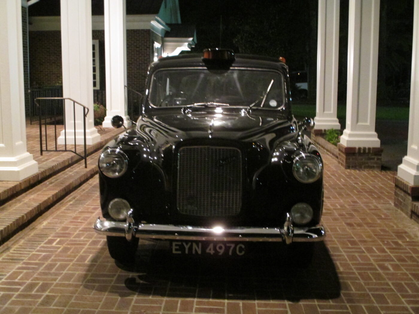 The taxi photographed head on at night under a portico, chrome bumper and round headlamps lit, London registration plate EYN 497C.