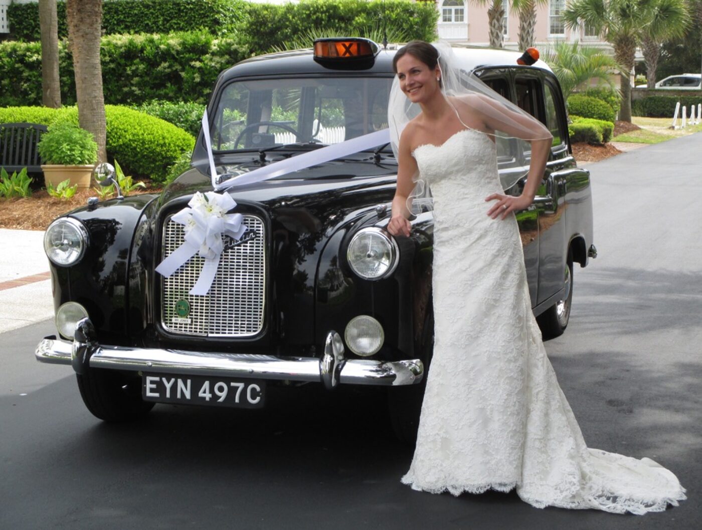 A bride standing at the front wing of the taxi, white ribbon and a spray of flowers tied to the radiator grille.